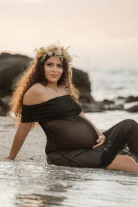 Future maman souriante en robe noire et couronne de fleurs, assise dans l’eau devant les rochers