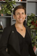 Une femme en veste noire sourit devant les étagères d’une bibliothèque et des plantes vertes