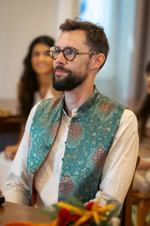 Le marié à lunettes en veste turquoise brodée regarde au loin, assis à table