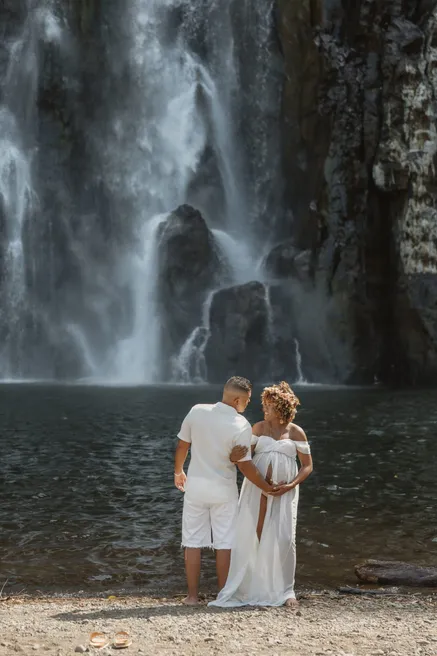 Un couple de dos face à une haute cascade, main dans la main au bord du bassin