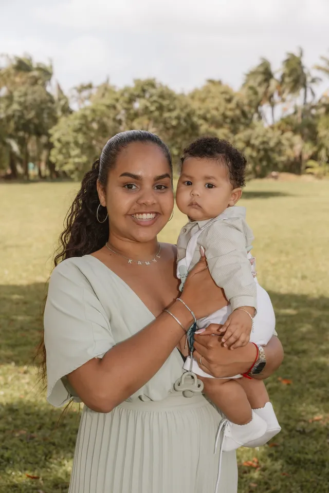 Une maman souriante tient son bébé en blanc dans ses bras, sur une pelouse bordée d’arbres