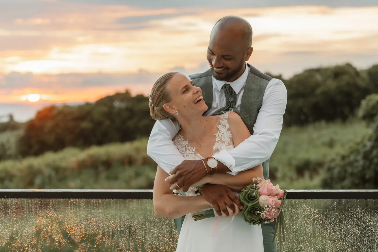 Le marié enlace par-derrière la mariée qui rit, devant un ciel de fin de journée