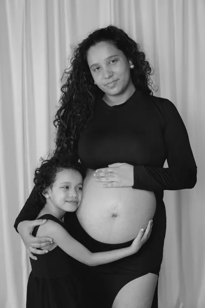 En noir et blanc, future maman en noir et sa fille qui l’enlace, devant un rideau clair