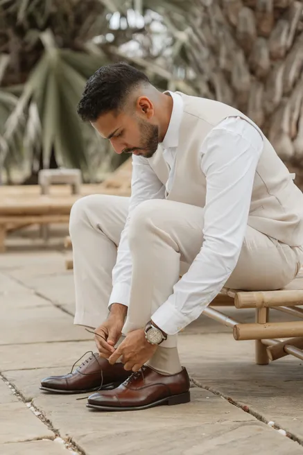 Le marié en gilet beige lace ses chaussures de cuir, assis sur un banc de bambou