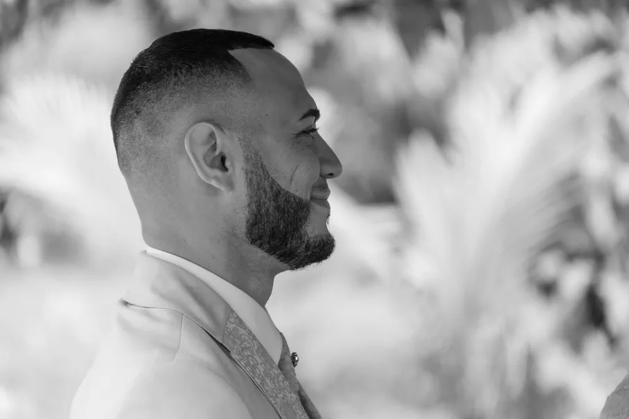 En noir et blanc, le marié barbu de profil, en costume clair, devant des palmes floues