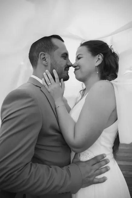 En noir et blanc, les mariés nez contre nez sous un voilage, la mariée souriante