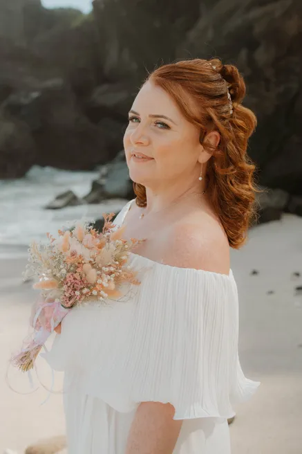 La mariée aux cheveux roux sur une plage, robe à épaules dénudées et bouquet de fleurs séchées