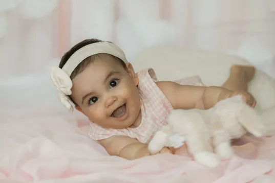 Un bébé allongé sur le ventre rit, bandeau à fleur blanche sur la tête, sur un drap rose