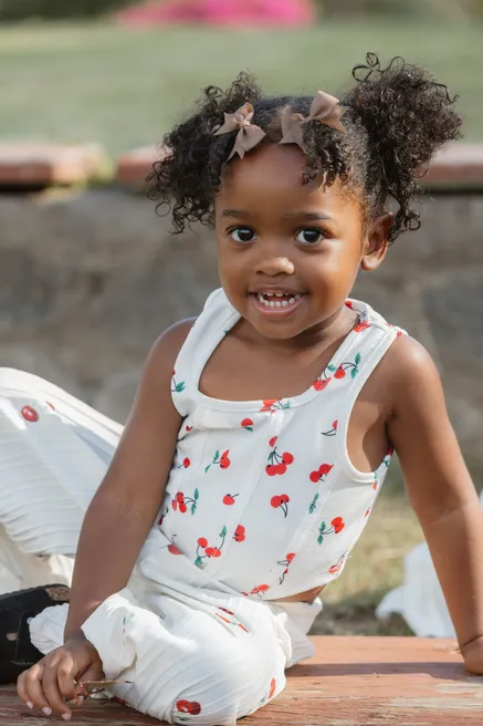 Une petite fille aux couettes bouclées sourit, assise dans un ensemble blanc à cerises