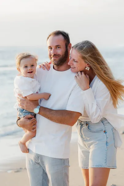 Un couple sourit sur la plage, le père porte contre lui leur bébé en salopette blanche