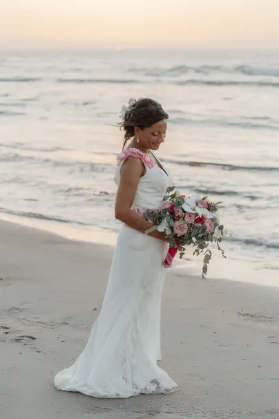 La mariée sur le sable, bouquet rose à la main, regarde les vagues à l’heure dorée