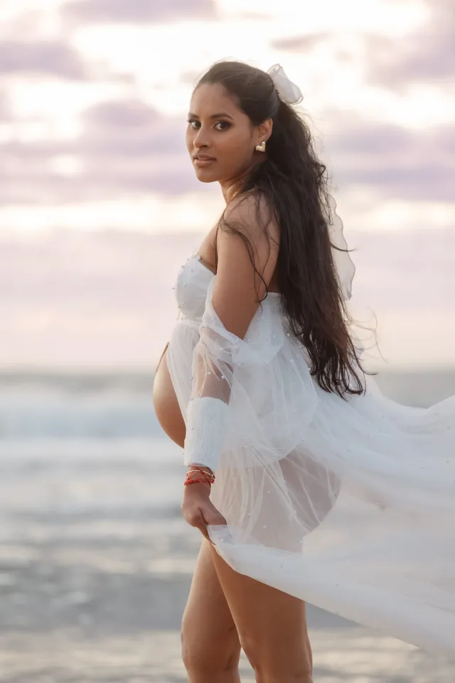 Future maman de dos qui se retourne, voile de tulle blanc porté par le vent, ciel mauve