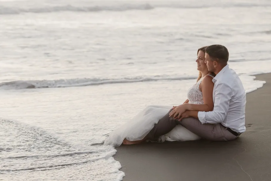 Les mariés assis sur le sable noir au bord des vagues, robe étalée dans l’écume