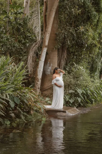 Future maman en robe blanche au pied d’un grand arbre, au bord d’un bassin en pleine végétation