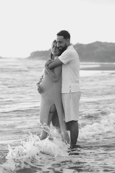 En noir et blanc, un couple enlacé dans l’écume, la future maman en robe longue