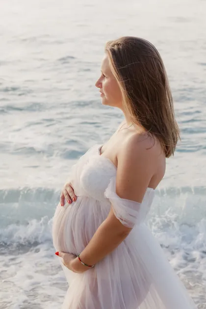 Future maman en robe de tulle, épaules nues, de profil face à la mer, mains sur le ventre