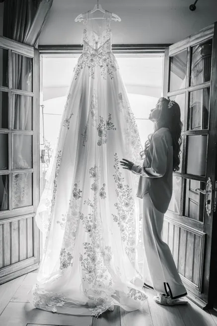 En noir et blanc, la robe de mariée suspendue à une porte-fenêtre, la mariée à côté