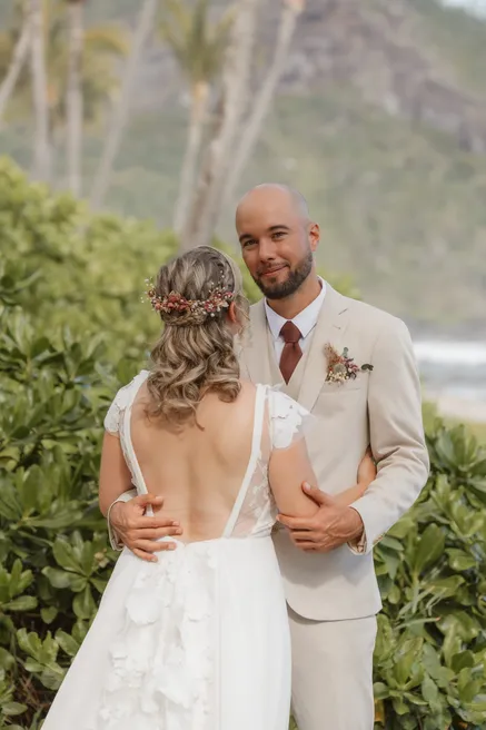 La mariée de dos, robe au dos nu, enlace le marié en costume beige dans la verdure