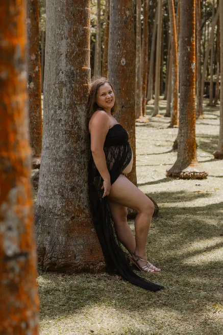 Future maman adossée à un tronc, longue robe noire, dans une forêt de troncs élancés