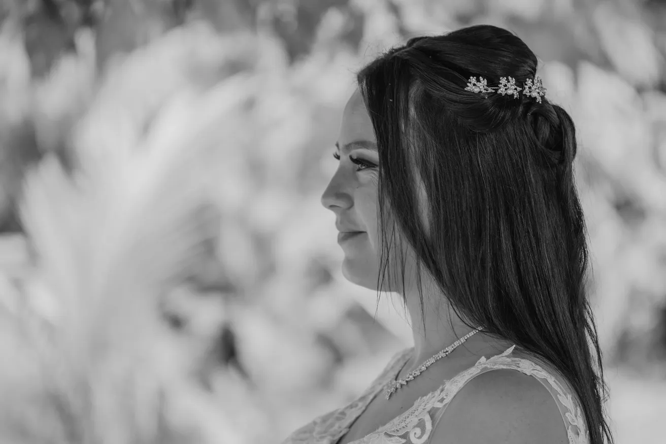 En noir et blanc, la mariée de profil, cheveux longs ornés d’un bijou, devant un feuillage flou