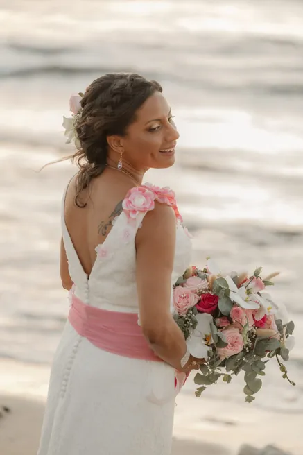 La mariée de dos regarde par-dessus l’épaule, ceinture rose et bouquet, devant la mer