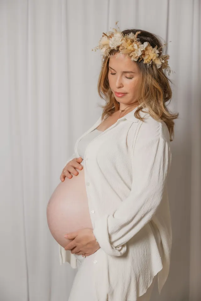 Future maman en chemise blanche et couronne de fleurs séchées, les mains sous le ventre, devant un fond clair