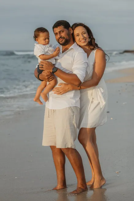 Un couple en blanc sourit pieds nus sur la plage, le père porte leur petit garçon