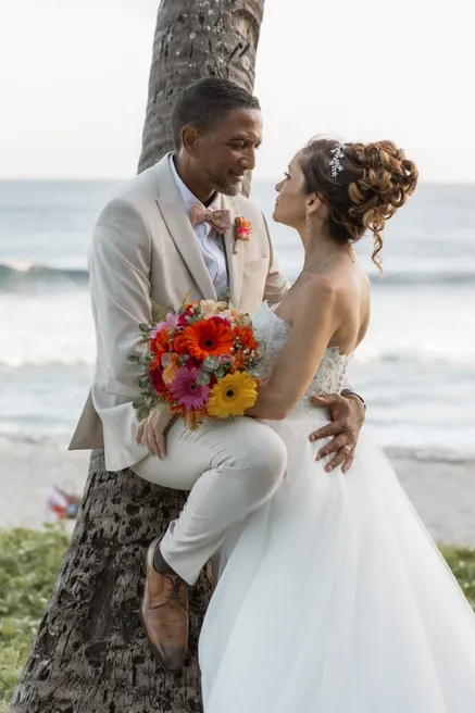 Les mariés enlacés contre un palmier face à la mer, bouquet de gerberas à la main