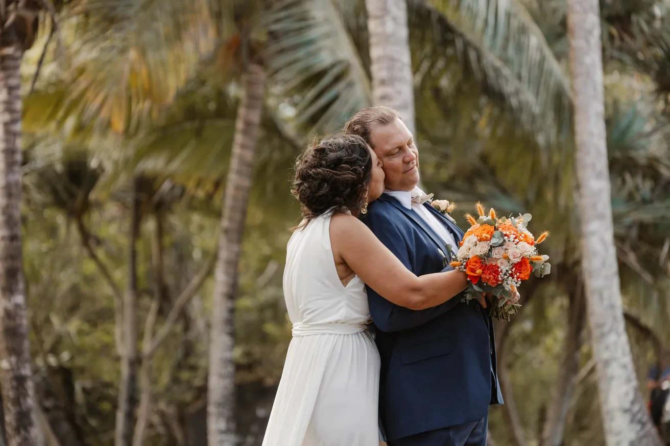 Les mariés enlacés entre les troncs de cocotiers, la mariée tient un bouquet orange