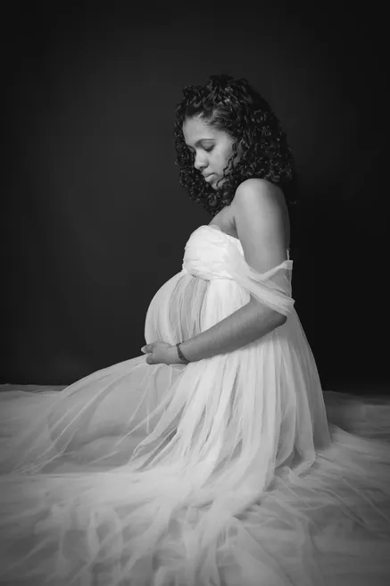En noir et blanc, future maman assise en studio, tête baissée, robe de tulle étalée