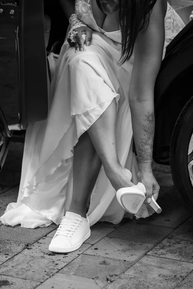 En noir et blanc, assise dans la voiture, la mariée enfile ses baskets blanches, un escarpin à la main