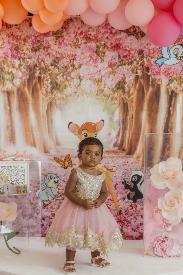Une petite fille en robe rose et dorée, debout devant un décor de fleurs roses et d’arbres