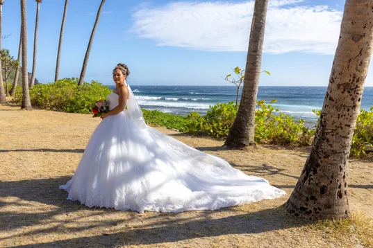 La mariée en robe à longue traîne de tulle, sous les cocotiers face à l’océan