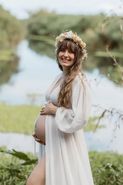 Future maman debout au bord d’une rivière, longue robe blanche ouverte et couronne de fleurs