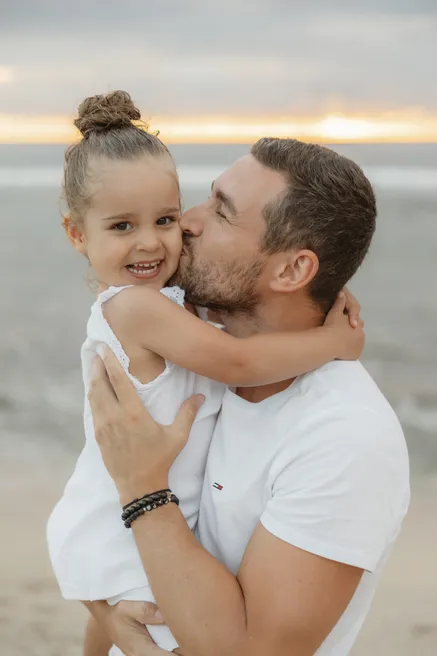 Une petite fille au chignon sourit, enlacée par son père qui l’embrasse sur la joue
