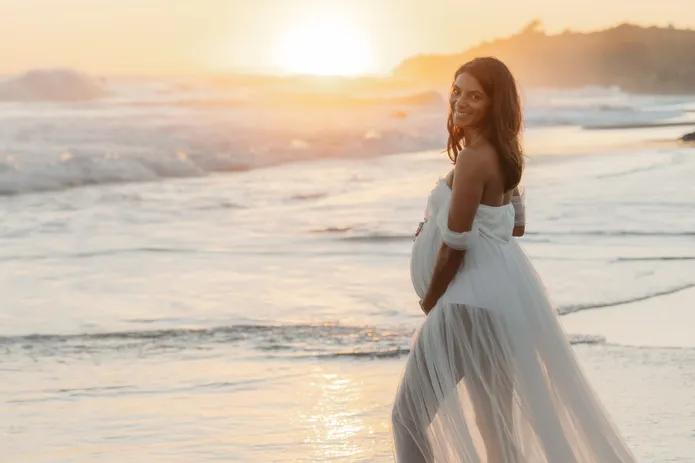 Future maman en robe de tulle blanc se retourne en souriant dans l’écume, au soleil couchant