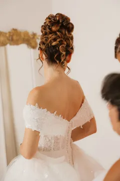 La mariée de dos, chignon bouclé et robe aux épaules dénudées, face à un miroir