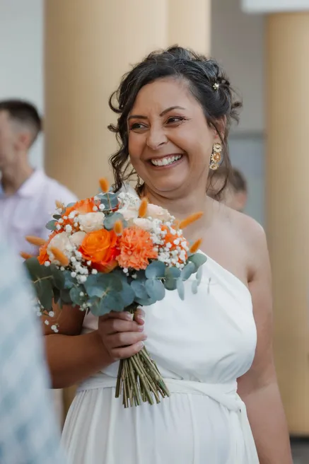 La mariée rit aux éclats, robe blanche asymétrique et bouquet de roses orange à la main