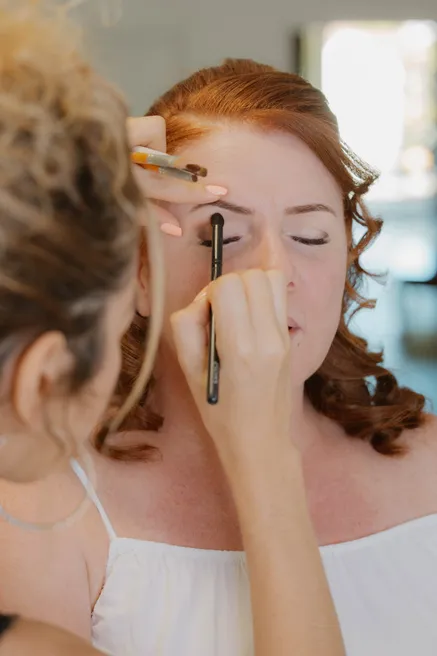La mariée ferme les yeux pendant que la maquilleuse trace le contour de ses sourcils