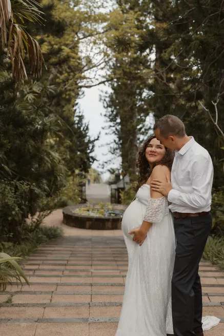 Un couple enlacé au bout d’une allée bordée d’arbres, la future maman en robe blanche