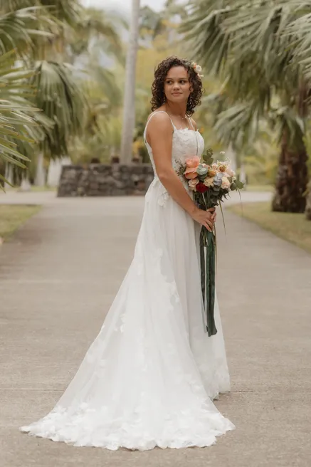La mariée seule dans une allée de palmiers, bouquet pêche à la main, longue robe de dentelle