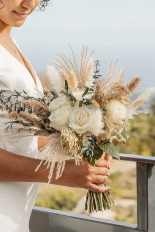 La mariée tient un bouquet de fleurs séchées et de roses blanches, accoudée à un balcon