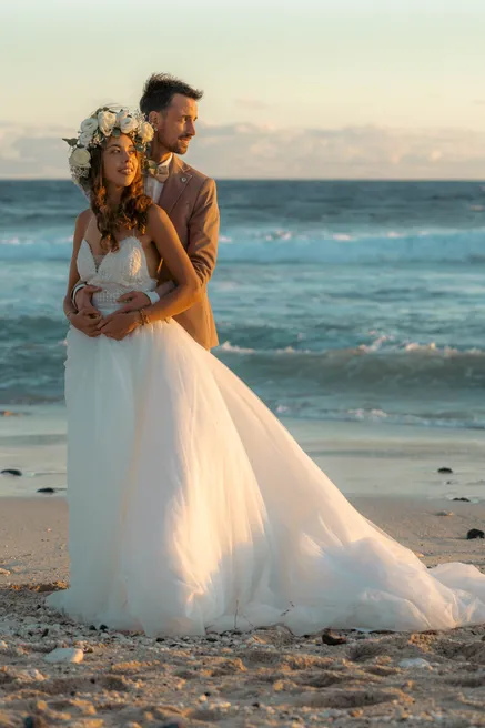 Le marié enlace la mariée en couronne de fleurs sur la plage, lumière dorée