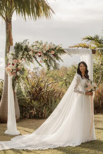 La mariée seule sous l’arche fleurie, traîne et voile étalés sur l’herbe