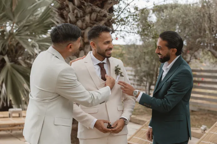 Deux témoins ajustent la boutonnière du marié en costume blanc, tous trois souriants
