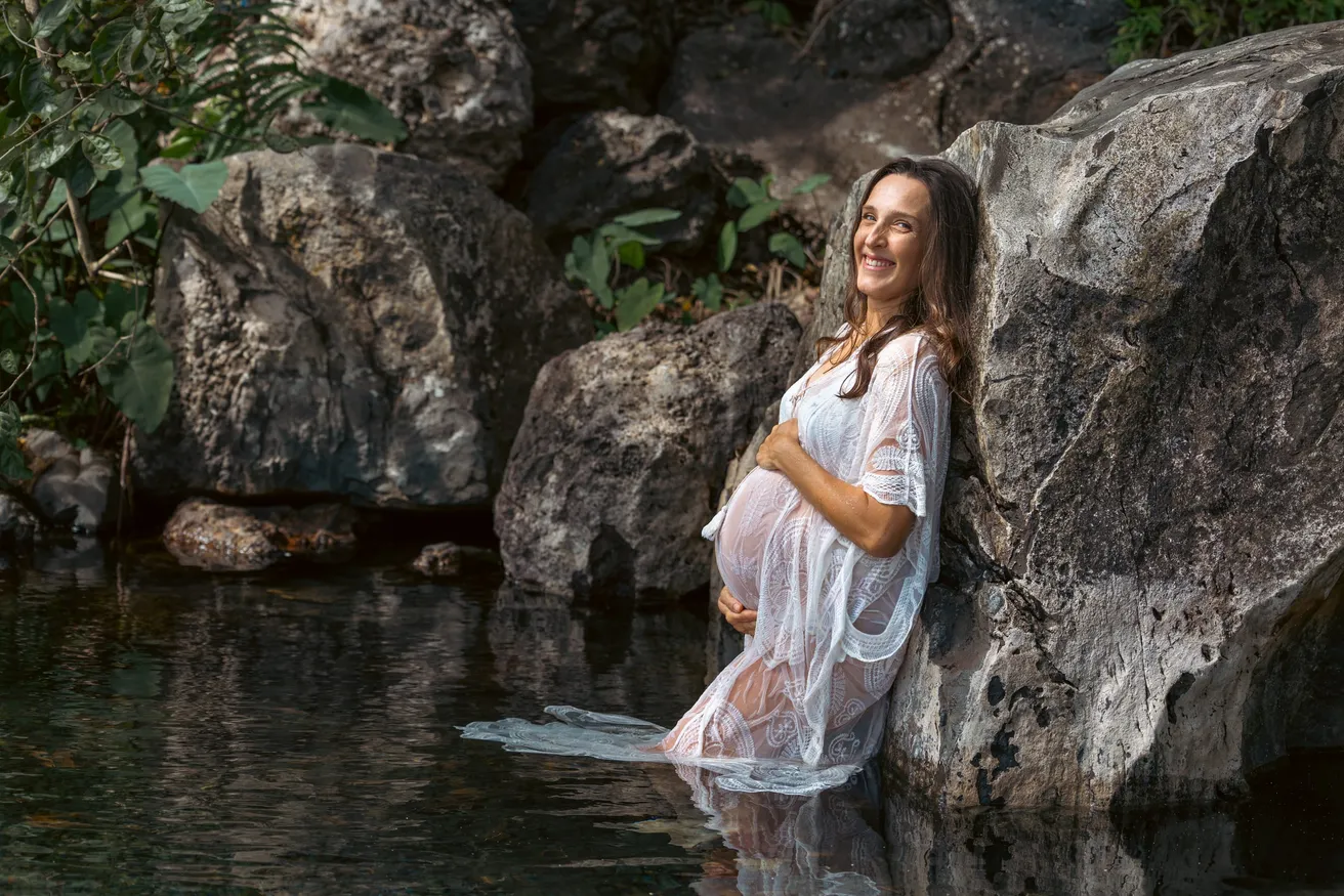 Future maman adossée aux rochers d’un bassin, en robe de dentelle blanche