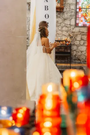 La mariée en voile et robe à traîne dans une église de pierre, vue entre des lumignons colorés