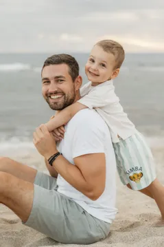 Un père rit, assis sur le sable, son petit garçon accroché à son dos