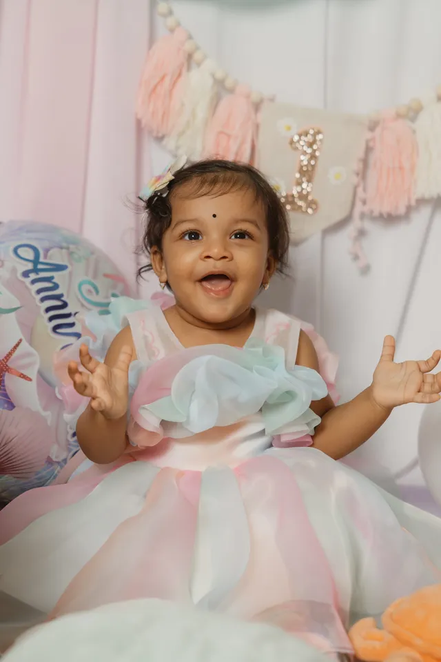 Une petite fille en robe pastel lève les bras en riant, un ballon d’anniversaire derrière elle