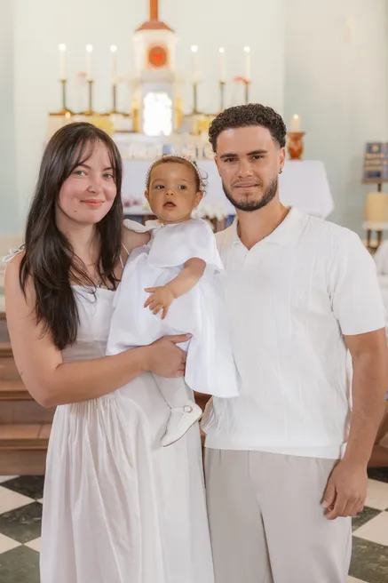 Un jeune couple en blanc tient son bébé à l’église, devant l’autel